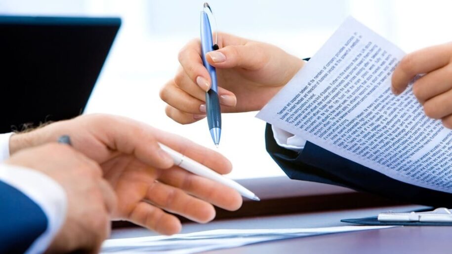 Firma de documentos. Manos de mujer sujetan una hoja de documento y bolígrafo. Mano izquierda de un hombre sujeta un bolígrafo blanco que señala a algún punto.