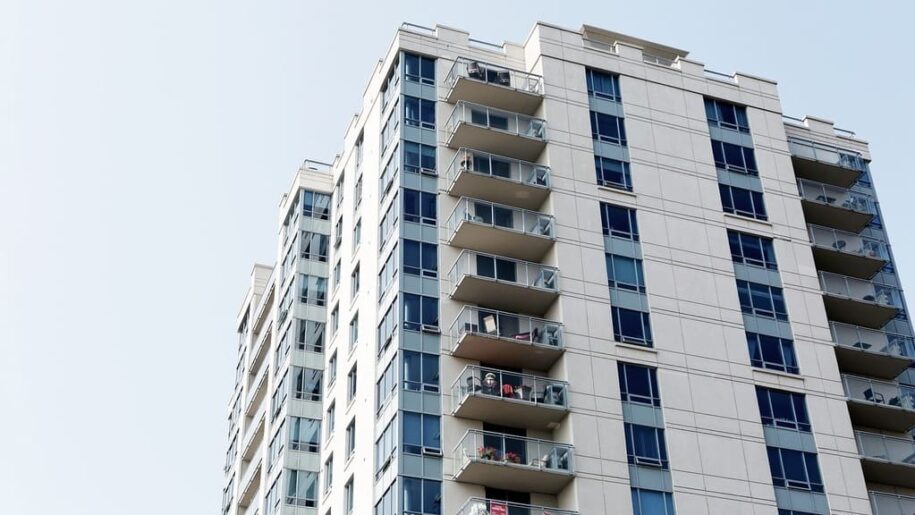 Edificio de más de 10 plantas, con fachadas que combinan balcones y numerosas ventanas. El edificio es color hueso. Cielo azul muy luminoso