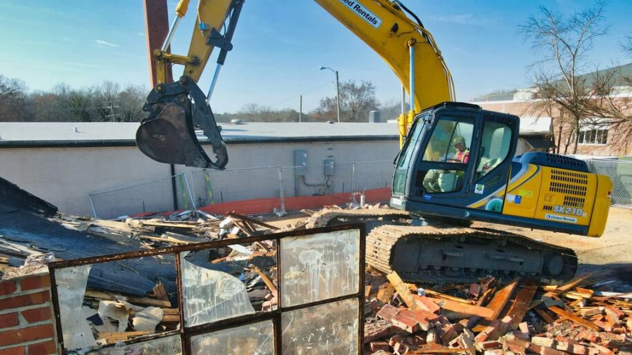 Tractor amarillo en proceso de demolición de una construcción. Resto de ventanas en primer término. Cielo azul al fondo.