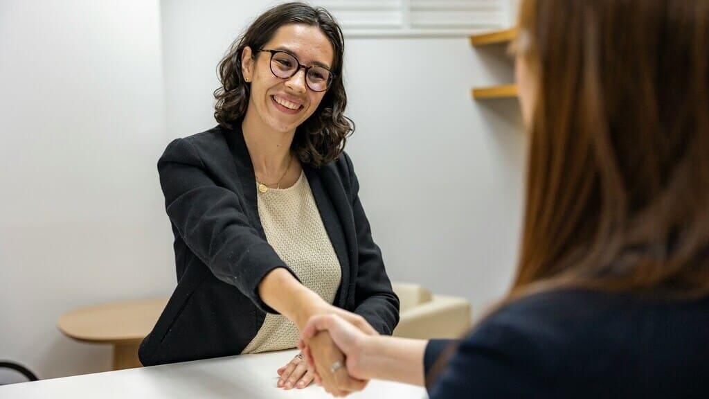 mujer joven morena con gafas, saluda dando la mano a otra mujer, de la que sólo se ve el brazo. Especialista en herencias, insolvencia y derecho urbanístico